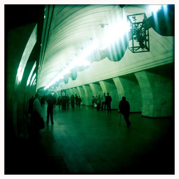 Green-tinted Hipstamatic photograph of a Moscow Metro station — arched marble ceiling, fluorescent lights, commuters silhouetted on the platform