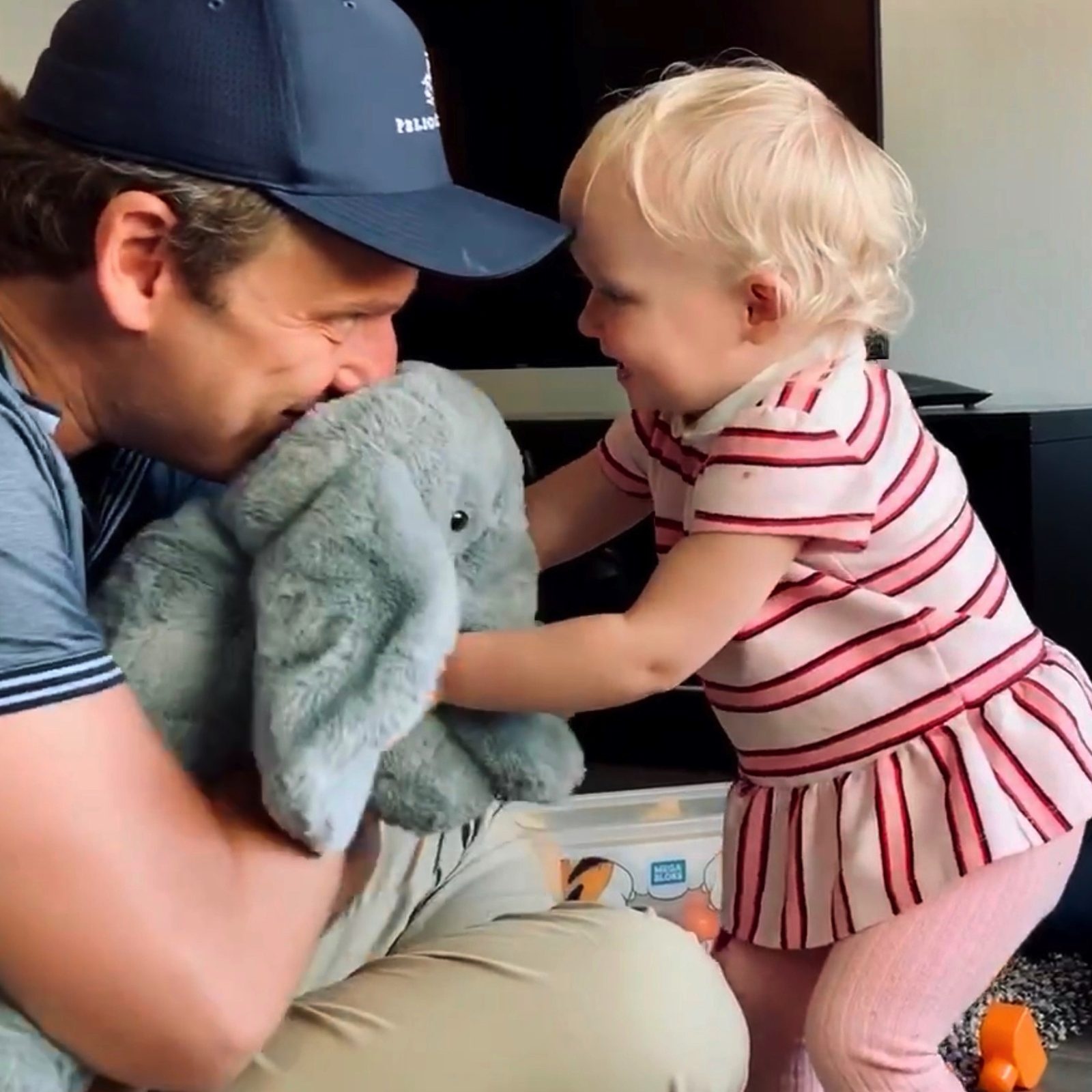 Steve Russell playing with toddler Evie during a supervised visit. He holds a stuffed grey elephant while Evie, in a red-and-white striped dress, reaches toward him laughing. Both are on the floor, face to face. Pelican Hill cap. Joy.