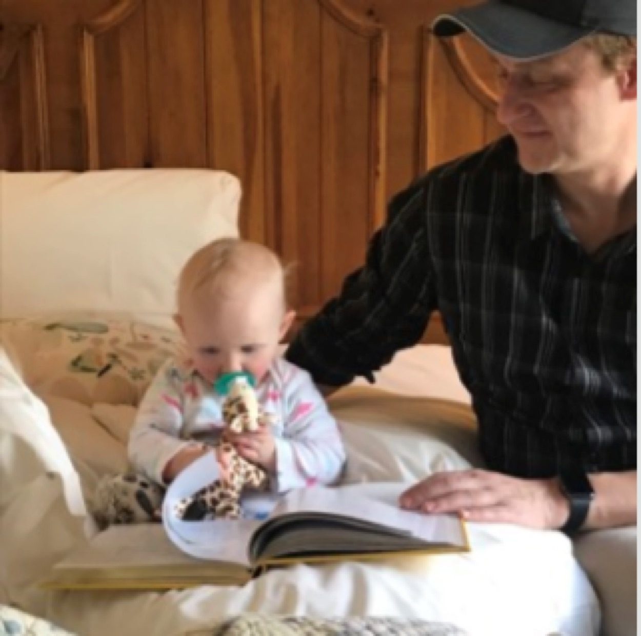 Steve reading to baby Evie on ornate bed at the Chappaqua rental — dark plaid shirt, cap, pacifier, giraffe, open book between them