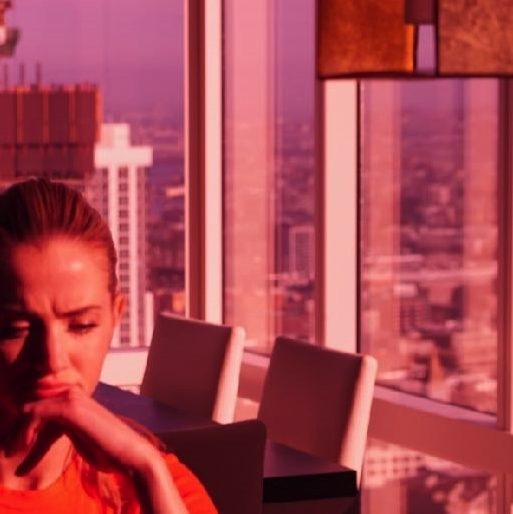 Tara Walsh in a high-rise San Francisco apartment at sunset — pink light through floor-to-ceiling windows, city skyline behind her, hands clasped, looking down