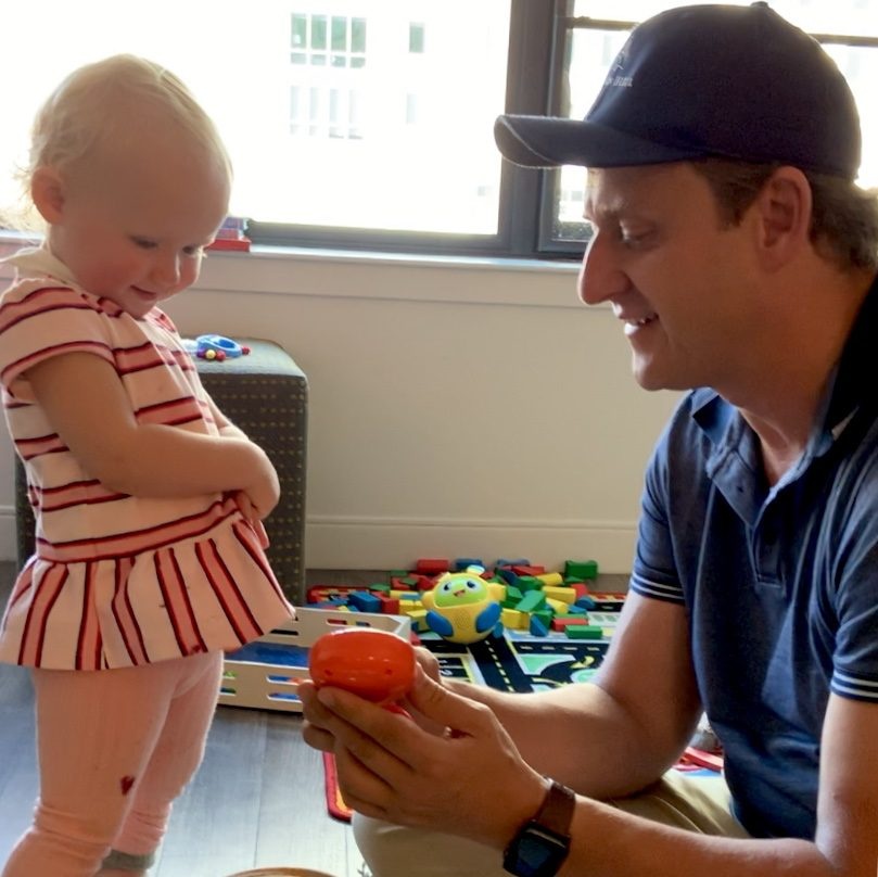 Steve Russell playing with toddler Evie — she stands in a red-striped dress, he kneels in a blue polo and baseball cap, offering her an orange ball, colorful toys on the floor